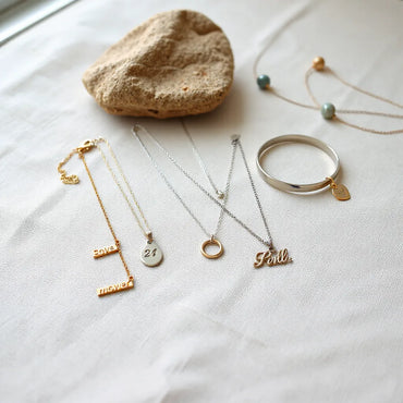 Assorted engraved necklaces and bracelets arranged on neutral fabric under soft natural light